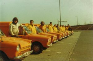 Eine Reihe von Männern in gelben Jacken, die zum RUWE-Team gehören, steht neben einer Reihe orangefarbener Transporter, die auf einer gepflasterten Fläche, möglicherweise auf einem Parkplatz, geparkt sind. Der Himmel ist klar, und im Hintergrund sind Straßenschilder und Straßenlaternen zu sehen.