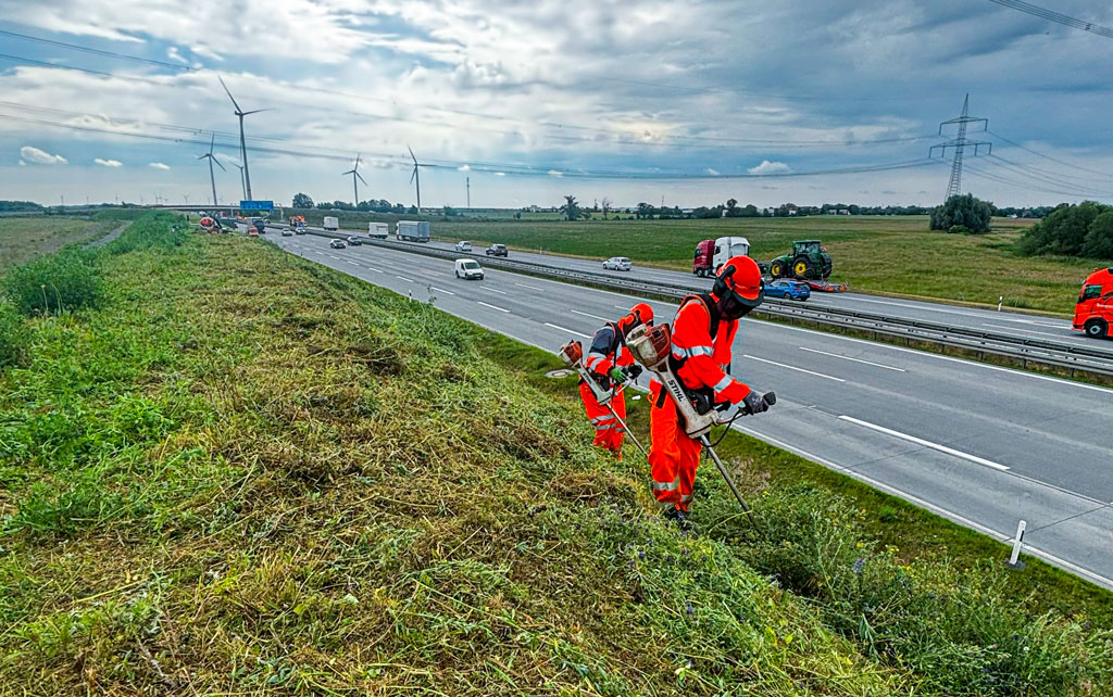 Zwei Arbeiter in orangefarbener Schutzausrüstung der RUWE Gruppe schneiden mit Rasentrimmern Gras auf der Böschung neben der mehrspurigen Autobahn Kappgraben Nord. Im Hintergrund sind Windräder, Stromleitungen und landwirtschaftliche Felder unter einem bewölkten Himmel zu sehen.