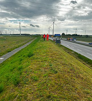 Zwei Personen in RUWE Warnkleidung laufen auf einem Grasdamm neben einer befahrenen Straße. Im Hintergrund sind mehrere Windkraftanlagen unter einem wolkigen Himmel zu sehen. Parallel zur Straße verlaufen Stromleitungen, die die Infrastrukturkompetenz der RUWE Gruppe unterstreichen.